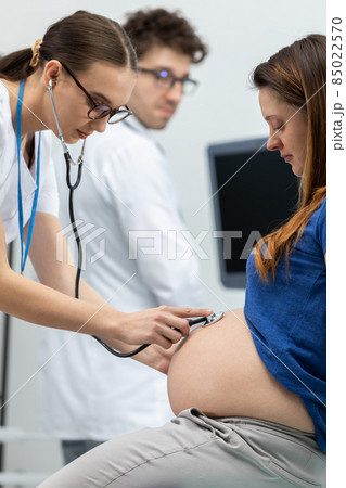 A lady doctor auscultates the abdomen of a pregnant woman with a stethoscope. In the background, a doctor is standing next to an ultrasound machine 85022570