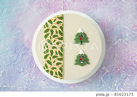 Beautiful and delicious white designer Christmas cake on the table with winter frosty pink and blue patterns with beautiful smiling Christmas trees in the form of chocolate cookies - top view 85023175