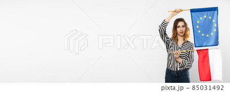 Young girl with the flag of the European Union and Poland. The student is holding two flags in her hand. Woman with flags on white background. Panoramic frame. Young girl with the flag of the European Union and Poland. The student is holding two flags in her hand. Woman with flags on white background. Panoramic frame. 85031492