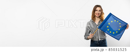 Panoramic frame. Young attractive girl with the flag of the European Union. A student holds the European Union flag in her hand. Woman with flag on white background. Isolated from the background. Panoramic frame. Young attractive girl with the flag of the European Union. A student holds the European Union flag in her hand. Woman with flag on white background. Isolated from the background. 85031525