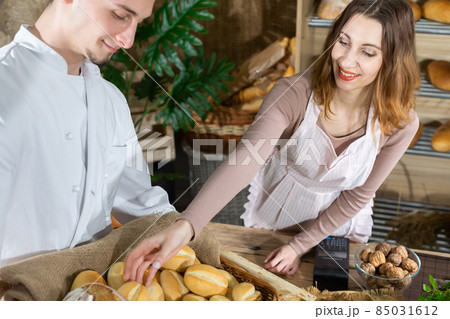 As fresh bread is delivered by a handsome baker, the attractive saleswoman offers one bun. Family bakery shop with multi-generation traditions. As fresh bread is delivered by a handsome baker, the attractive saleswoman offers one bun. Family bakery shop with multi-generation traditions. 85031612