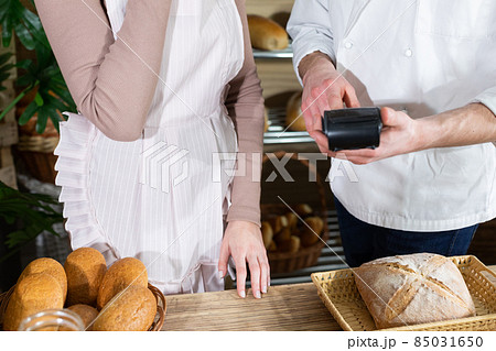 A baker teaches his new saleswoman how to operate a payment machine. Family bakery shop with multi-generation traditions. A baker teaches his new saleswoman how to operate a payment machine. Family bakery shop with multi-generation traditions. 85031650