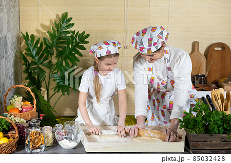 Mom and daughter roll out pizza dough with a wooden rolling pin on one board in the kitchen. Mother and baby do the dough together in the kitchen. Mom and daughter roll out pizza dough with a wooden rolling pin on one board in the kitchen. Mother and baby do the dough together in the kitchen. 85032428