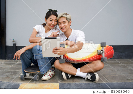Young asian couple sit resting after skateboarding, take a selfie. Young man in a white T-shirt holding tablet computer while the woman pointing finger at his face. Young asian couple sit resting after skateboarding, take a selfie. Young man in a white T-shirt holding tablet computer while the woman pointing finger at his face. 85033046