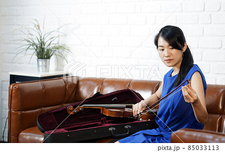 Young asian violinist in blue dress takes care of the instrument after practice session in living room. Rubbing rosin on the horse hair of the violin bow. Young asian violinist in blue dress takes care of the instrument after practice session in living room. Rubbing rosin on the horse hair of the violin bow. 85033113