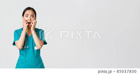 Covid-19, healthcare workers and preventing virus concept. Scared insecure asian female doctor, nurse in scrubs starts to panic from fear, biting fingernails and looking horrified, white background 85037830