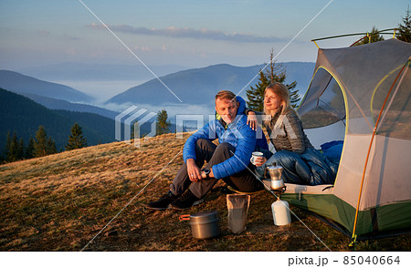 Man and woman resting at campsite in the mountains. Breakfast of two campers outdoors on background of beautiful mountain hills covered green forests. Concept of hiking, camping and relationships. 85040664
