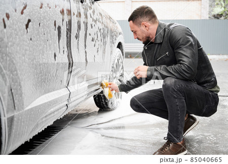 Young man washing car on carwash station outdoor. Handsome driver cleaning automobile, using sponge and soap. 85040665