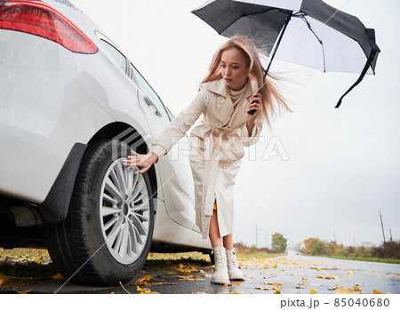 Beautiful woman standing on a road near her white car with punctured car tire. Female driver looking on wheel, holding umbrella. 85040680