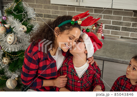 Happy mom with antler hoop, her adorable son in Santa hat kissing her on cheek, enjoying spending happy time together in winter Christmas holidays. Happy family relationships, childhood, motherhood 85043846