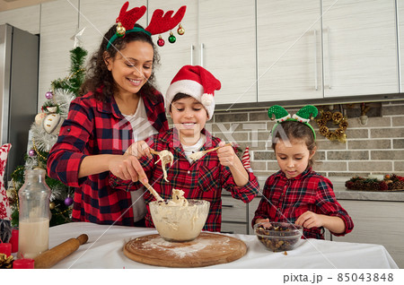 Charming woman, mother teaches children how to make dough for Christmas baking. Adorable European children cook with their mother in kitchen. Concept to share a happy time during Christmas holidays 85043848