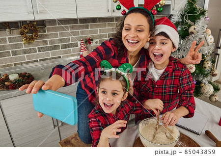 Christmas mood at home kitchen. Beautiful mom and her adorable children- boy and girl smiling toothy smile looking at web camera of smartphone, making selfie, recording video while cooking together 85043859
