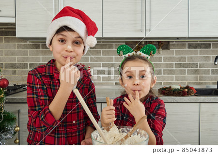 Cheerful beautiful children in checkered red and green clothes looking at the camera holding a finger in their mouth, tasting the dough they have prepared for the Christmas cake Cheerful beautiful children in checkered red and green clothes looking at the camera holding a finger in their mouth, tasting the dough they have prepared for the Christmas cake 85043873
