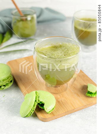 Matcha tea in a glass and green macaroon cake on a wooden bamboo tray. Close-up on a gray background. Matcha tea in a glass and green macaroon cake on a wooden bamboo tray. Close-up on a gray background. 85050844