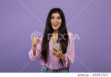Yes, great news. Surprised armenian woman celebrating win, using smartphone and pointing at gadget, purple background 85061507