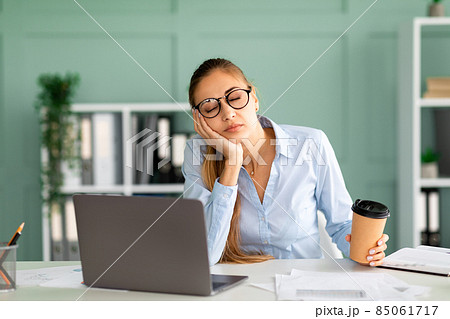 Young businessman tired of computer work sleeping at workplace, holding coffee cup in hand Young businessman tired of computer work sleeping at workplace, holding coffee cup in hand 85061717