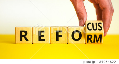 Refocus and reform symbol. Businessman turns a wooden cube and changes the word refocus to reform. Beautiful yellow table, white background. Business refocus and reform concept. Copy space. 85064822