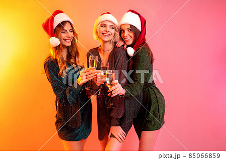 Three happy girls in Santa Claus hats holds glasses of champagne having fun. New Year party 85066859