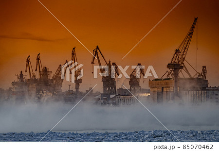 The construction of nuclear icebreakers at magic sunset, cranes of of the Baltic shipyard in a frosty winter day, steam over the Neva river, smooth surface of the river, sky of orange color 85070462