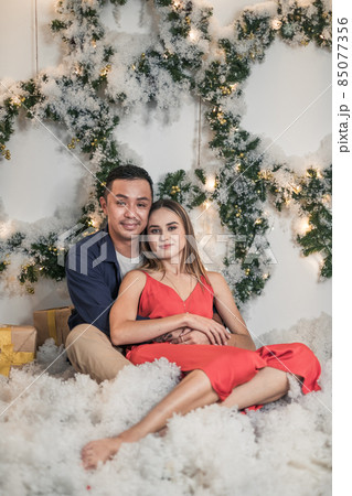Christmas studio family shoot. New Year's photo of happy couple. Christmas wreaths and artificial snow. Man and woman cuddle. Christmas studio family shoot. New Year's photo of happy couple. Christmas wreaths and artificial snow. Man and woman cuddle. 85077356