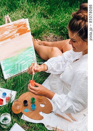 Artist painting on the easel outdoors in the garden. Open air outdoor art workshop. Draw on the canvas with brush and palette sitting on the grass during a picnic in a park Artist painting on the easel outdoors in the garden. Open air outdoor art workshop. Draw on the canvas with brush and palette sitting on the grass during a picnic in a park 85078086