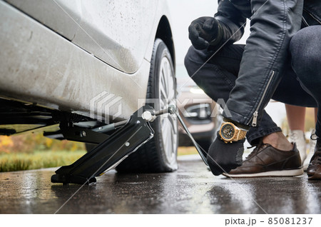 Close up of male hands raising vehicle with car jack before changing flat tire. Young man with watch on his wrist using special device for lifting car while repairing woman vehicle on the road. 85081237