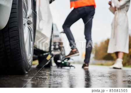 Close up of automobile on rainy road with female driver and roadside assistance worker on blurred background. Male auto mechanic repairing woman car on the street. Focus on care wheel. 85081505