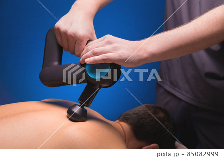 Close-up of a professional male masseur stimulates the back muscles of a male patient in a dark spa room for massage. Percussion mechanical effect on overstrained muscles Close-up of a professional male masseur stimulates the back muscles of a male patient in a dark spa room for massage. Percussion mechanical effect on overstrained muscles 85082999