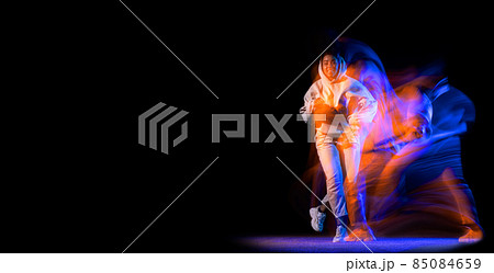 Sportive young girl in white costume dancing hip-hop dance isolated on dark background at dance hall in neon mixed light. Sportive young girl in white costume dancing hip-hop dance isolated on dark background at dance hall in neon mixed light. 85084659