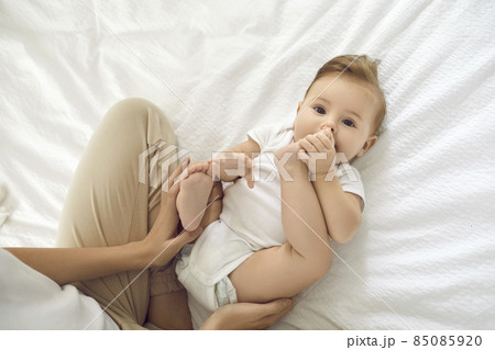 Portrait of cute charming six month old girl playing with her legs lying on her back on white cotton bed in bedroom. Baby in white bodysuit lying next to unrecognizable mother and looking at camera. 85085920