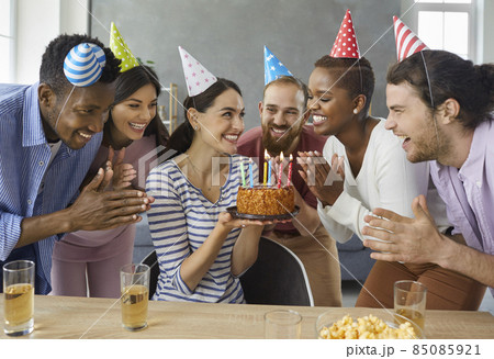 Group of cheerful young multiracial people in conical party hats applaud while the birthday girl makes a wish. Happy woman holding a birthday cake while celebrating at home with her best friends. 85085921
