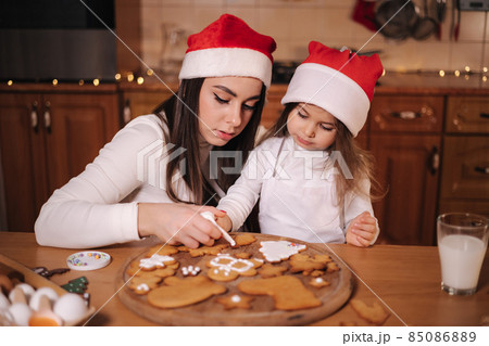 Mom in santas hat with her little doughter decorates gingerbread using glaze. Christmas and New Year traditions concept. Christmas bakery. Happy holidays Mom in santas hat with her little doughter decorates gingerbread using glaze. Christmas and New Year traditions concept. Christmas bakery. Happy holidays 85086889