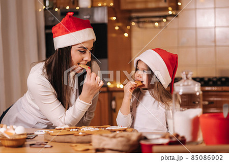 Adorable little girl and her mom in santa hat eating homemade gingerbread and laughing. Happy little kid preparing for Christmas. Background of fairy lights bokeh 85086902