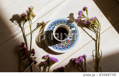 Turkish coffee in traditional blue porcelain cup at saucer on a white wooden floor top view. Contrasting shadows. Hot espresso drink in a morning light flatly. A composition of dried purple flowers.  85088749