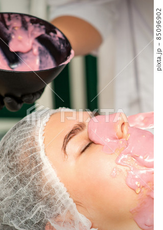 The cosmetologist applying an alginate mask to the face of a young woman in a beauty salon. 85096902