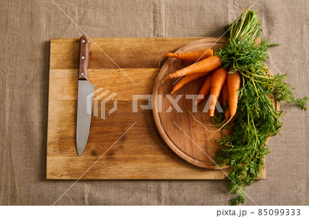 Flat lay of a big bunch of fresh carrots with green tops and a kitchen knife on a wooden cutting board on a linen tablecloth. Food background of seasonal raw vegetables Flat lay of a big bunch of fresh carrots with green tops and a kitchen knife on a wooden cutting board on a linen tablecloth. Food background of seasonal raw vegetables 85099333