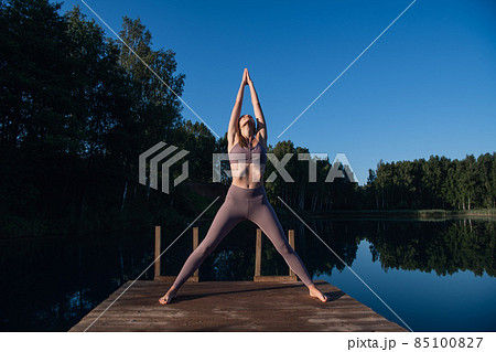 Young woman by the lake practicing yoga moves on wooden platform. Pretty young woman exercising in nature, healthy lifestyle young people positive vibes. Young woman by the lake practicing yoga moves on wooden platform. Pretty young woman exercising in nature, healthy lifestyle young people positive vibes. 85100827