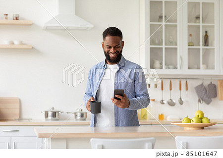 Cheerful young black male with cup of coffee in hand in minimalist kitchen interior reads news on smartphone Cheerful young black male with cup of coffee in hand in minimalist kitchen interior reads news on smartphone 85101647