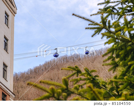 Two blue lifts ride a cable car against the backdrop of mountains with bare trees. View through the spruce branches Two blue lifts ride a cable car against the backdrop of mountains with bare trees. View through the spruce branches 85104550