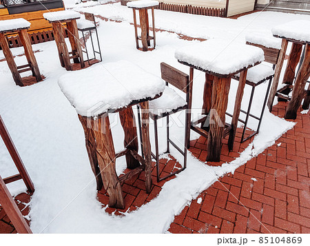 High wooden tables and bar stools of an outdoor cafe stand in a row in winter covered with snow 85104869