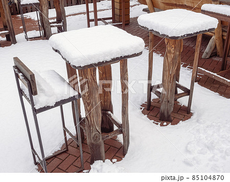 Tall wooden tables and bar stools of an outdoor cafe stand covered with snow in winter 85104870