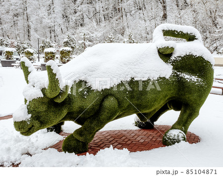 Green statue of a bull made of artificial grass stands covered with snow on the street near the forest 85104871