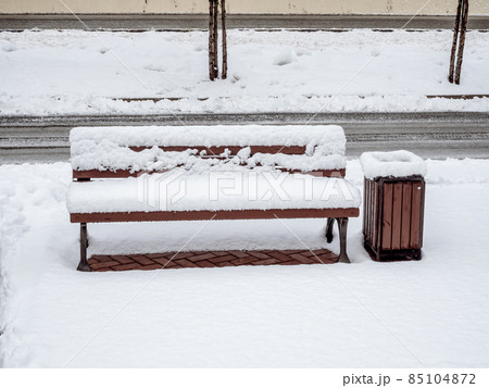 A bench and a trash can stand outside strewn with fresh white snow 85104872