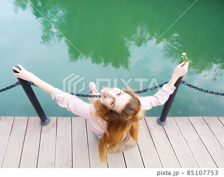 A smiling girl sits on a wooden bridge over the lake holding on to iron pillars. Top view 85107753