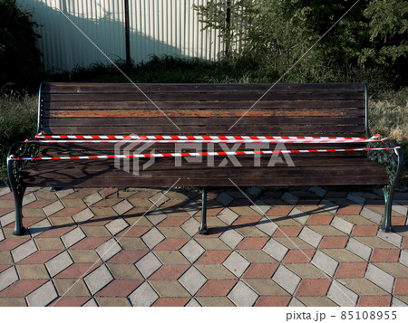Wooden painted bench covered with protective tape stands on paving slabs on a sunny day Wooden painted bench covered with protective tape stands on paving slabs on a sunny day 85108955