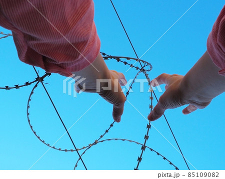 Hands under the barbed wire reach up against the blue sky 85109082