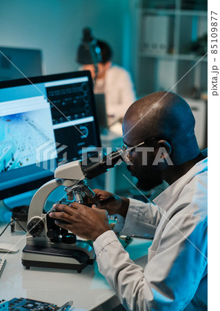 Young man of African ethnicity sitting by workplace and looking in microscope 85109877