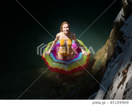Smiling girl in a multicolored swimming circle stands in the water in the rays of the sun in a shady gorge of canyons Smiling girl in a multicolored swimming circle stands in the water in the rays of the sun in a shady gorge of canyons 85109909