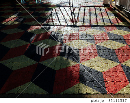 Terrain with paving slabs with a red-black geometric pattern and a falling shadow from trees and a fence Terrain with paving slabs with a red-black geometric pattern and a falling shadow from trees and a fence 85111557