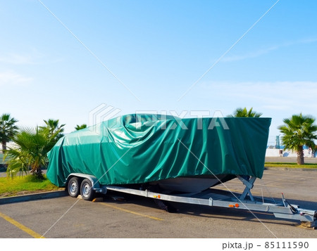 A motor boat covered with a turquoise cover stands on land under the open sky against the background of palm trees A motor boat covered with a turquoise cover stands on land under the open sky against the background of palm trees 85111590